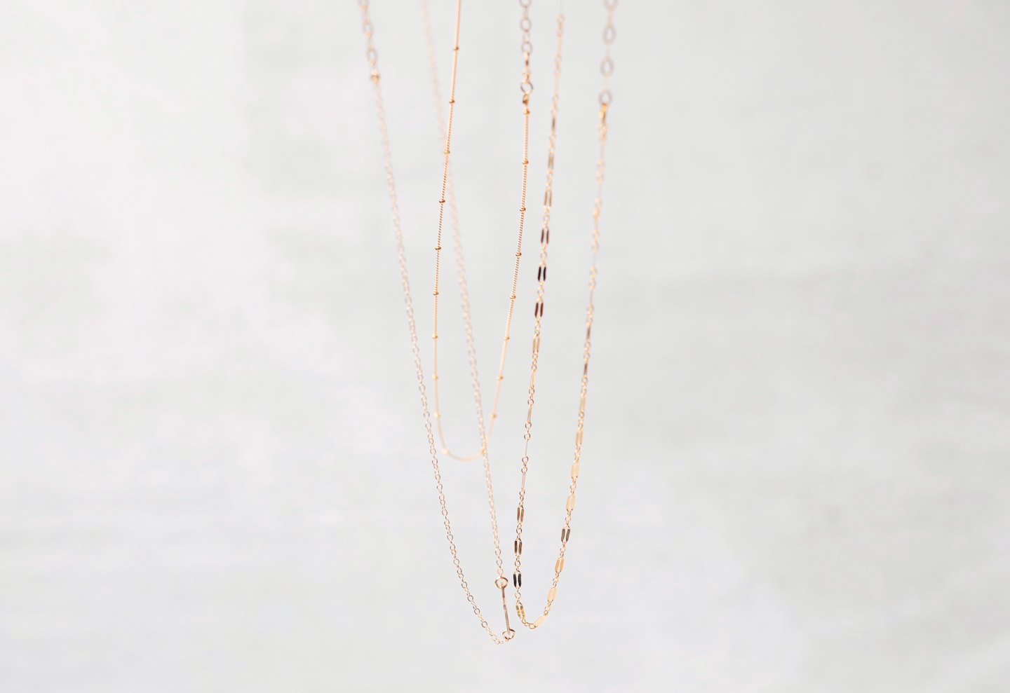 Three delicate gold necklaces hang vertically against a plain wall.